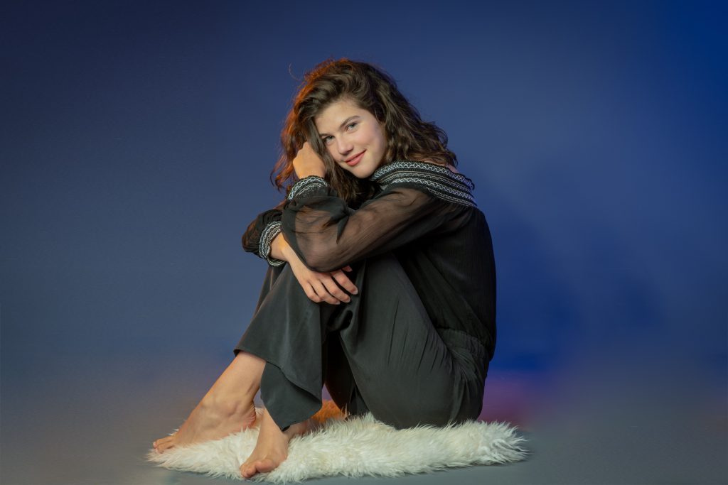 Jeune femme assise sur peau de mouton , songeuse. Fond dégradé bleu/gris. Photo Studio Polidori