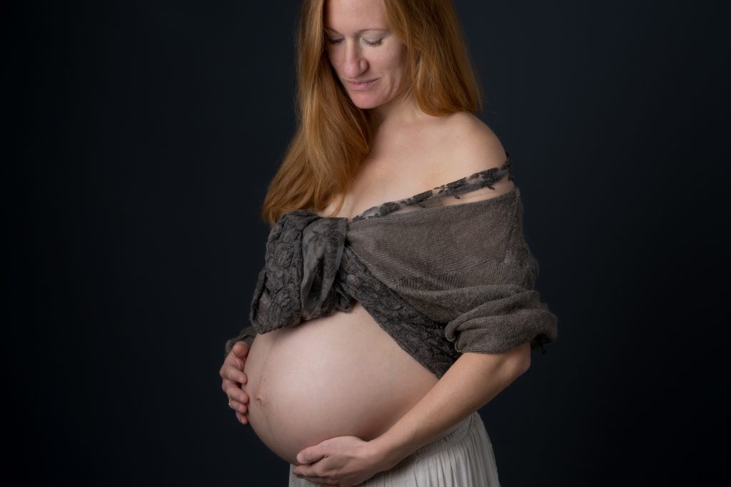 Femme enceinte type bohème rousse regardant son ventre sur fond noir. Photo Studio Polidori