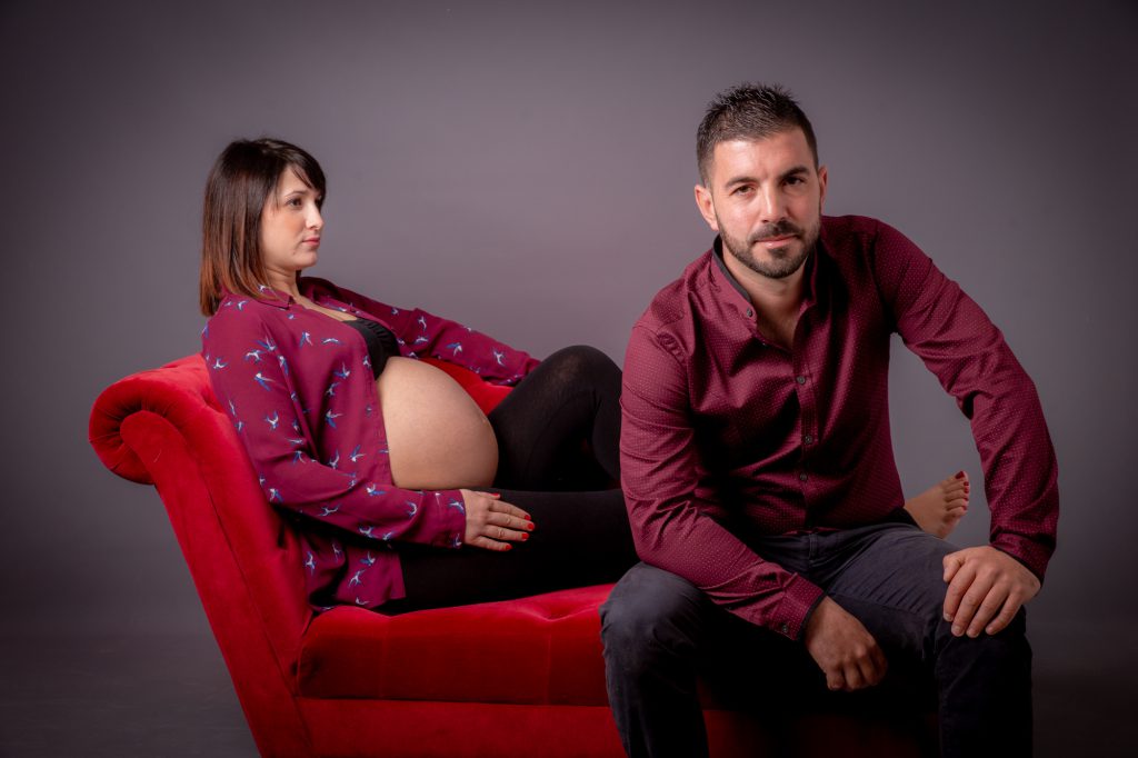 Futur papa assis avec madame enceinte (ventre rond) en arrière plan). Photo Studio Polidori