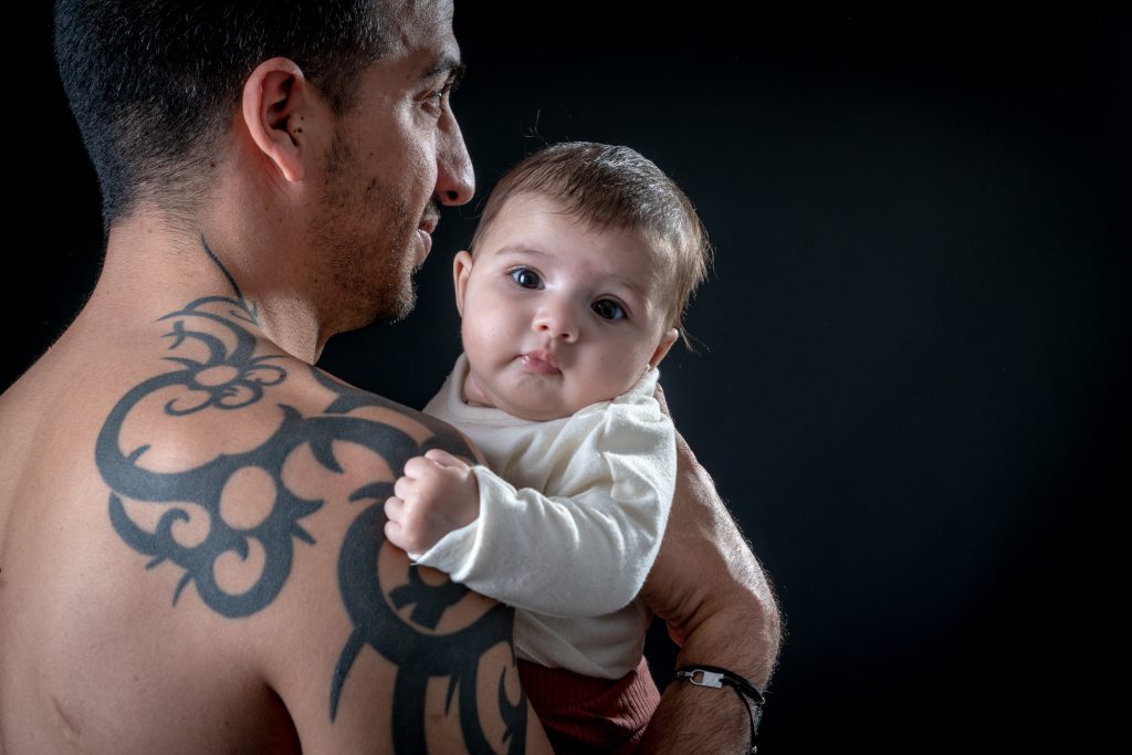 Bébé bien calé sur l'épaule de son papa, nous regarde, yeux grands ouverts. Photo Studio Polidori